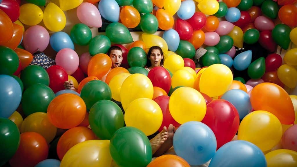 two adults a lamp and some other household items surrounded by balloons