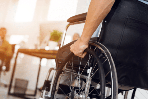 A-disabled-man-is-sitting-in-a-wheelchair.-He-holds-his-hands-on-the-wheel.-Nearby-are-his-colleagues