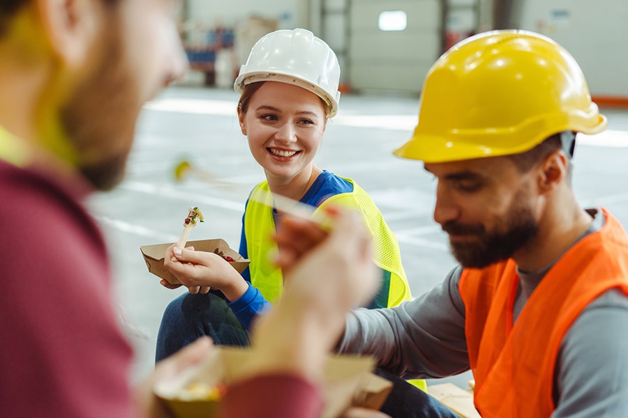 Group of workers, managers, wearing hard hats and vests having lunch, eating, smiling, talking during break at work. Concept of lunch, food, communication