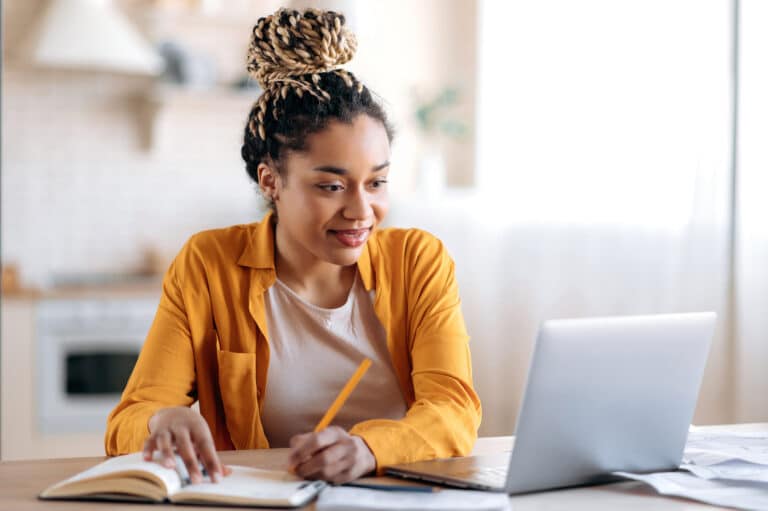 Focused african american female student studying remotely from home, using a laptop, taking notes on notepad during online lesson, e-learning concept, smiling