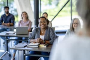 A class of mature students sit in class together as they listen attentively to the teacher. They are each dressed casually and have laptops and books out in front of them as they take notes during the teaching.