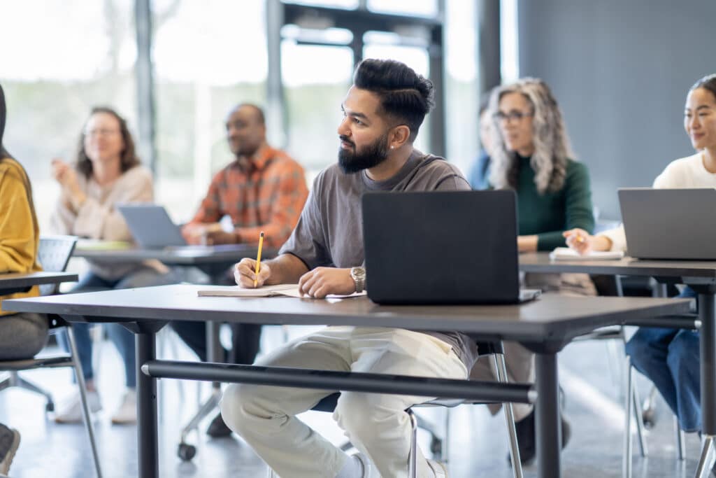 A large group of mature adults are seen sitting at desk with wither laptops and books open as they study during a continuing education class. They are each dressed casually and are listening attentively.