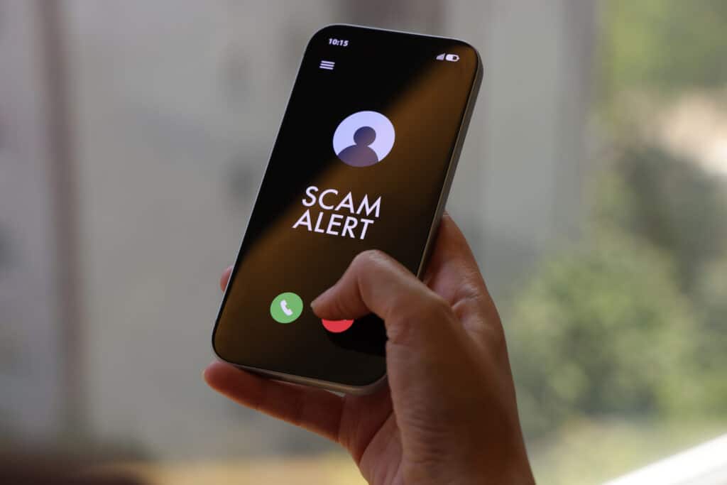 A person holds a smartphone displaying a “Scam Alert” message on an incoming call screen. The dark interface clearly indicates a fraud warning, conveying concerns about cybersecurity, mobile fraud, and digital safety. The indoor environment and close-up composition highlight the immediacy and relevance of online threat awareness in everyday mobile use.
