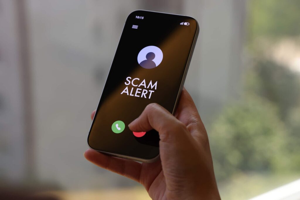 A person holds a smartphone displaying a “Scam Alert” message on an incoming call screen. The dark interface clearly indicates a fraud warning, conveying concerns about cybersecurity, mobile fraud, and digital safety. The indoor environment and close-up composition highlight the immediacy and relevance of online threat awareness in everyday mobile use.