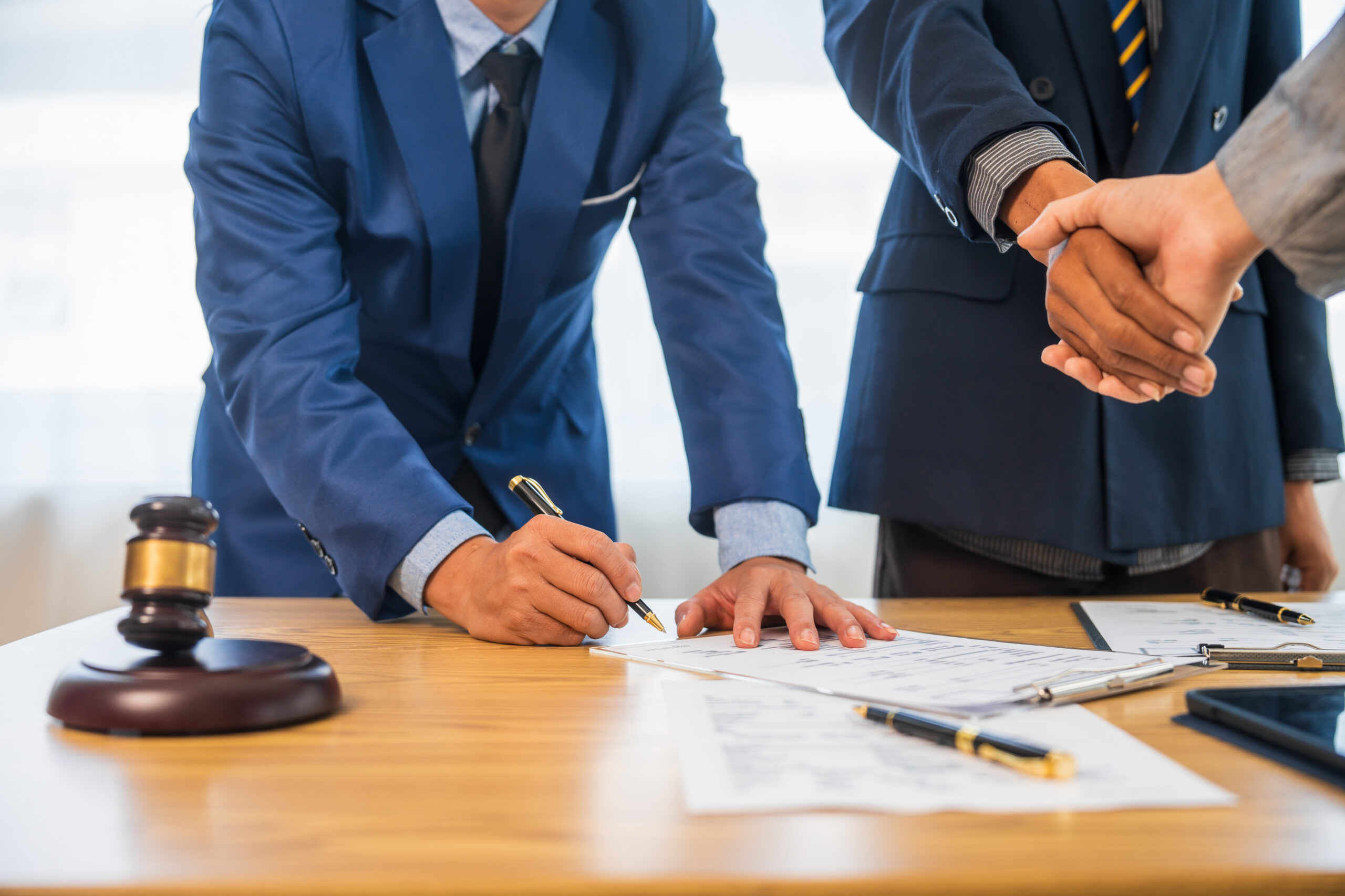 A team meeting of business people and a lawyer in formal suits is taking place at a desk, discussing a contract and various aspects of the law and litigation.
